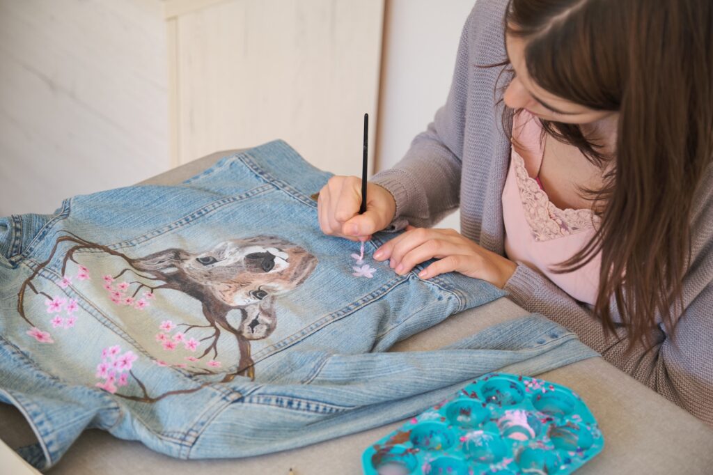 Woman hand painting with acrylic paints a deer and flowers in a denim jacket.