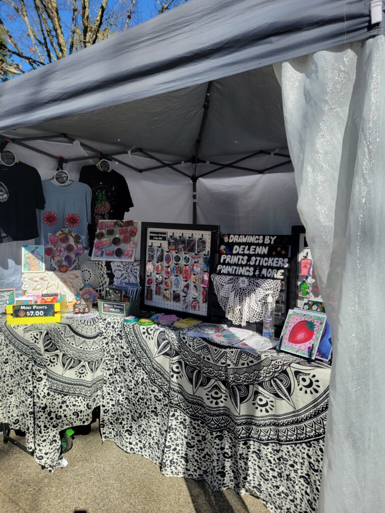 A Staurday Market Booth filled with original artwork. T-shirts hang in the back, an array of prints, and stickers are displayed on black and white table cloths.